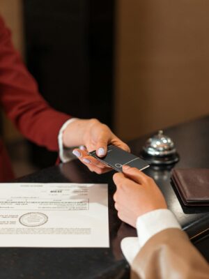 A hotel receptionist hands a key card to a guest over the counter, with documents visible.