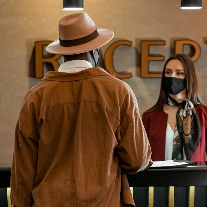 Man with luggage checking in at hotel reception. Staff wearing mask, indoors, travel theme.